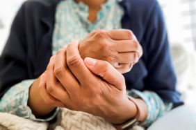 Close up of a woman's hands/ dry skin with a darker skin tone. The woman is wearing a patterned light blue blouse and a dark blue sweater; there is a watch on her wrist 