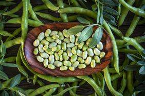 Wooden bowl of shelled raw lima beans (green) surrounded by green bean pods
