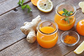Two jar-shaped shot glasses of wellness immunity juice made with ginger, citrus, turmeric, honey. The ingredients—whole ginger root, lemon slices, orange, turmeric powder—sit besides the glasses on a wood table.