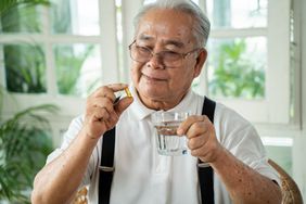 Older Asian man wearing a white polo and black suspenders sits at home, holding a fish oil pill and a glass of water. Windows and plants are behind him in the background.