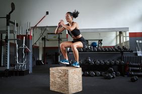 Young woman wearing black athletic clothes and blue sneakers is performing a box jump. In a gym setting, she is about to land in a squat position on top of a plywood square. 