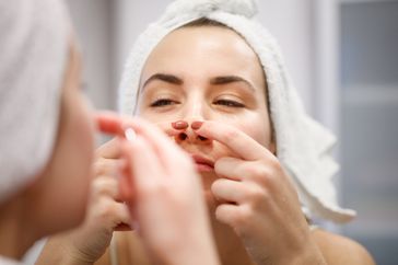 Young white woman wears a white towel on her head and examines her nose closely in the bathroom mirror