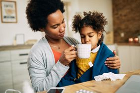 Parent gives tea to child on their lap who appears to have a cold.