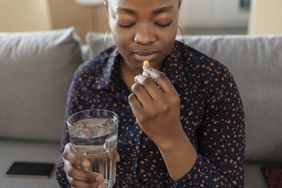 Young black woman wearing a patterned shirt is taking a supplement pill and holding a glass of water, sitting on a couch