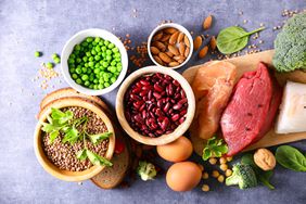 Overhead view: Raw chicken, steak, fish on a cutting board. Bowls of dry beans, peas, lentils. Almonds, eggs, chickpeas and broccoli.