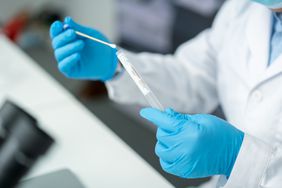 A pair of gloved hands holds a swab and test tube.