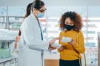 Pharmacist in a convenience store with her hair in a pony tail wears a mask and glasses and shows a customer, wearing a yellow sweater with black curly hair, also wearing a mask, a supplement.
