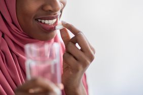 Close up of a black woman wearing a pink hijab taking a white supplement pill and holding a glass of water