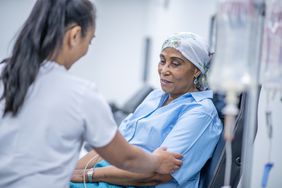 A Black woman wearing a headscarf undergoing chemotherapy treatment is being attended to by a female healthcare professional in a hospital setting