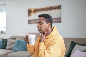 A man has his eyes closed and puts his hand up to his throat as he holds a mug