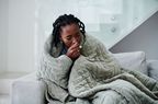 Woman sits on couch wrapped in blanket and coughing.