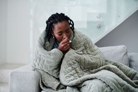 Woman sits on couch wrapped in blanket and coughing.