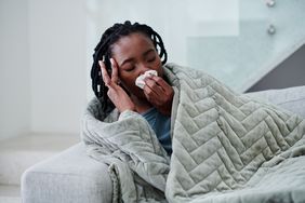 Person feeling sick, lying on couch in blanket with tissues.
