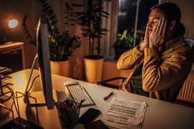 A man is tired as he puts his hands to his face and sits at a computer at nighttime