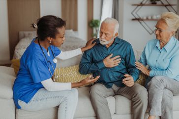 older man sitting alongside loved one and doctor on a couch touching his chest in pain