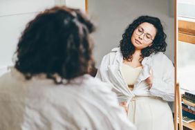 Young curly haired woman wearing a white outfit and round glasses looking at her body in the mirror