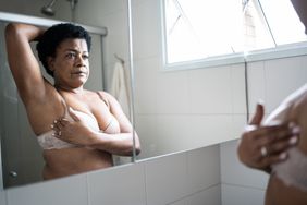Woman with short black hair standing in a bra gives herself a breast exam in the mirror.