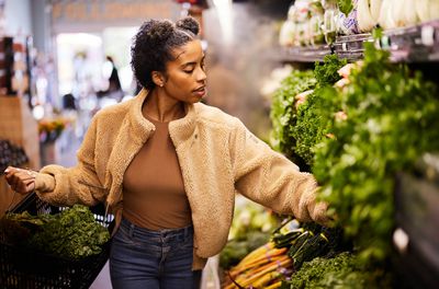 Young black woman wearing a fuzzy tan coat and jeans is shopping in the produce aisle at the grocery store