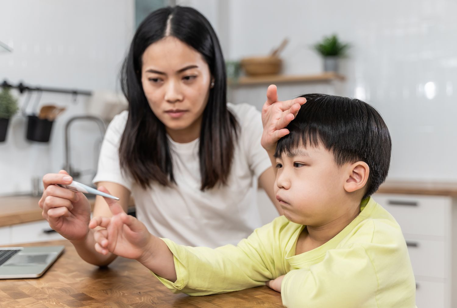 Portrait of Asian mother checking her sick sons temperature, looking at thermometer and touching forehead of her son.