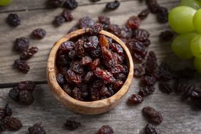 A small wooden bowl of raisins (dried grapes) on a wooden surface. More raisins are scattered around the bowl, and whole green grapes sit to the side