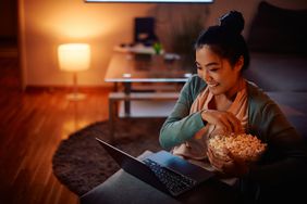 Young Asian woman sitting at home on her couch watching a movie on her laptop and eating a late-night snack of a bowl of popcorn