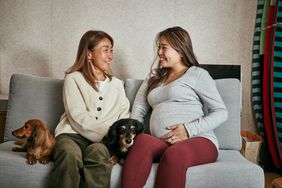 Pregnant person and mom sit on couch with two dogs.
