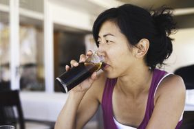 Asian woman sitting at a cafe drinking a tall class of cola soda with a lemon. She wears a purple tank top and has her hair tied back in a loose bun.