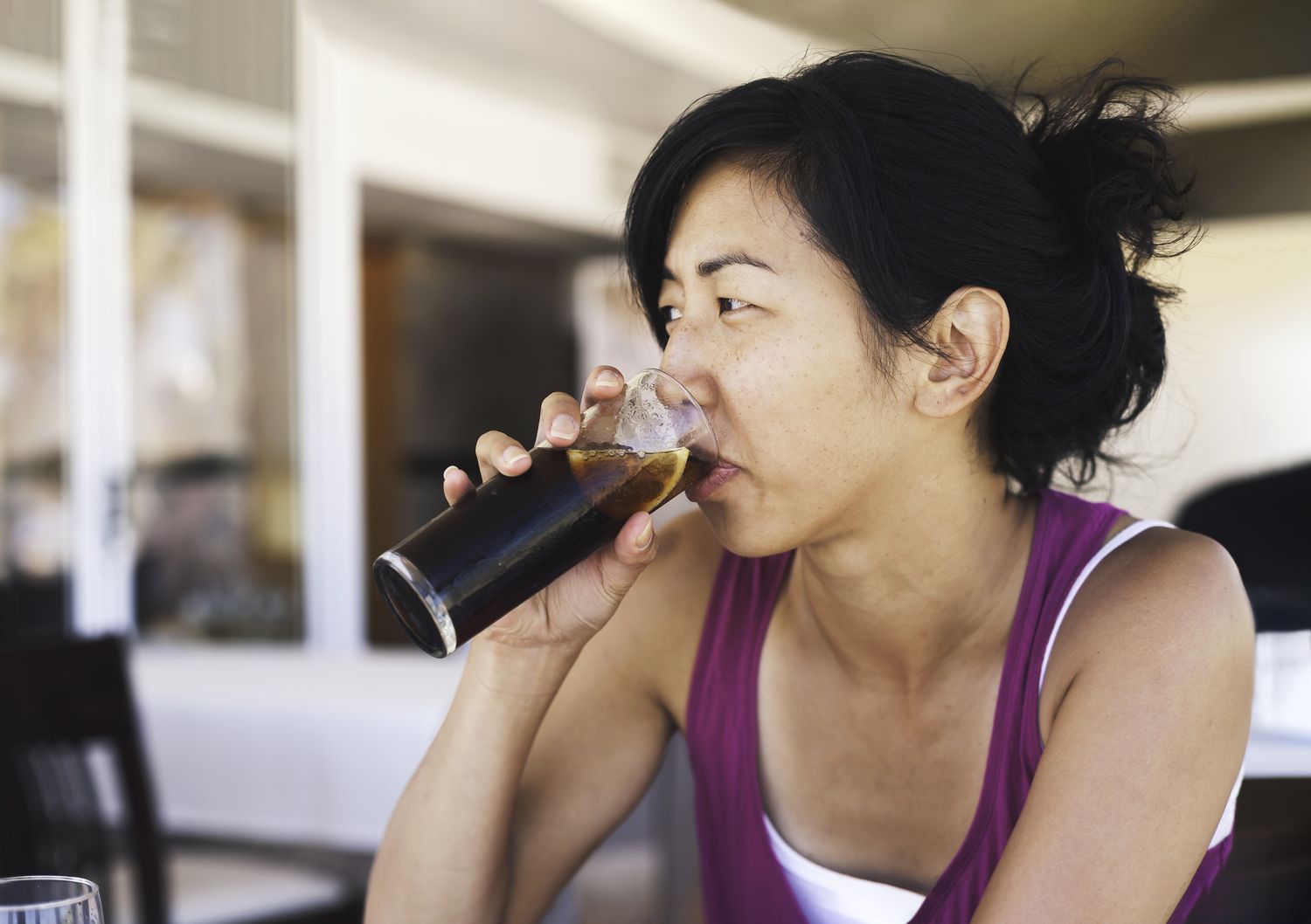 Asian woman sitting at a cafe drinking a tall class of cola soda with a lemon. She wears a purple tank top and has her hair tied back in a loose bun.