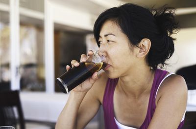 Asian woman sitting at a cafe drinking a tall class of cola soda with a lemon. She wears a purple tank top and has her hair tied back in a loose bun.