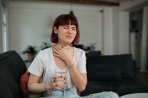 A young woman holds a hand up to her throat in pain as she holds a glass of water in the other hand