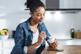 Black woman with brown curly hair pulled into a ponytail, wearing a jean button down shirt and tshirt, sits in her kitchen and eats a cup of yogurt, looking down at her smartphone on the table