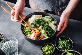 Person holding chopsticks over a bowl of vitamin k-rich foods: soybeans; herbs; leafy greens. The bowl also has salmon, cucumber, and rice