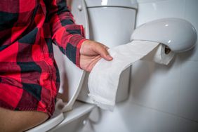 person with a red and black plaid shirt sitting on the toilet and grabbing toilet paper