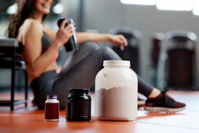 Three sizes of powder workout supplement containers sit on a gym floor. In the background, a woman wearing athletic clothes sits on the floor and is holding a shaker bottle.