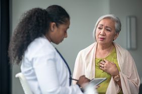 A senior woman shares her chest pain symptoms with her doctor at an appointment. The woman is dressed casually and holding her chest with her hand as she looks downward with a distraught expression. The doctor is sitting in front of her listening attentively.