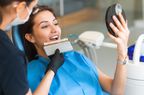A dentist holds up a row of veneers against a patient's mouth as the woman looks on in a mirror