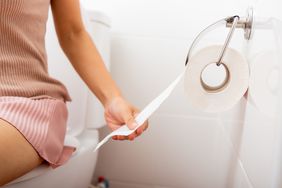 A person sits on the toilet bowl and pulls a roll of toilet paper from the wall