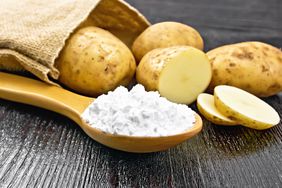 A wooden spoon holding potato starch (white powder), laid flat on a wooden surface. A sack of potatoes spills out behind the spoon; one potato is half sliced