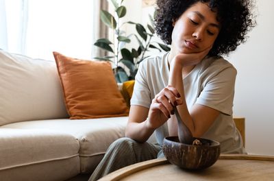Young multiracial woman eating breakfast feeling tired. She sits by a tan couch and wood coffee table, a plant is in the background.