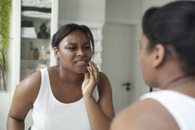 Black woman examines her mouth in the mirror; touching her jaw in pain from grinding her teeth all night