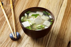 A bowl of miso soup (broth, tofu, green onion) and a pair of chopsticks resting on a small blue chopstick holder, on a wooden table