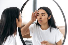 A woman puts a finger to her forehead as she inspects her face in the mirror