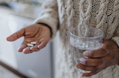 Close up of person's hands holding two supplement pills and a glass of water. The person is wearing a tan cable knit sweater and is standing in a kitchen