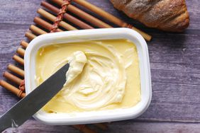 Overhead view of a tub of butter with a butter knife scooping up butter. A loaf of bread sits on the edge of the frame