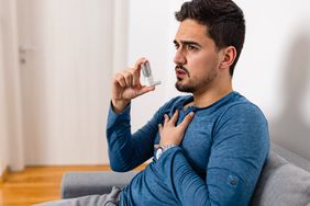 A man sits with one hand over his chest as he holds an inhaler