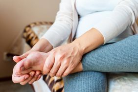 Pregnant woman wearing leggings and a cardigan is sitting at home with her foot propped up on her leg, massaging her feet and toes.