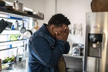 man in denim jacket standing in the kitchen holding hands over face experiencing sadness