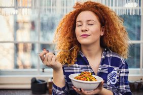 Woman with bright red, curly hair wearing a blue plaid shirt is eating a healthy meal in her kitchen