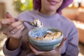 Young black woman wearing purple sweatshirt and bonnet, eating oatmeal with buts and fruit at home