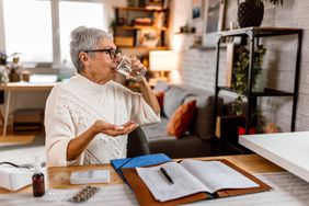 older woman in a white sweater sitting on a chair and drinking water as she takes a medication